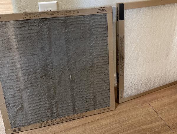 A dirty air filter sitting to the left of a clean air filter on top of a wood plank floor and leaning against a white wall