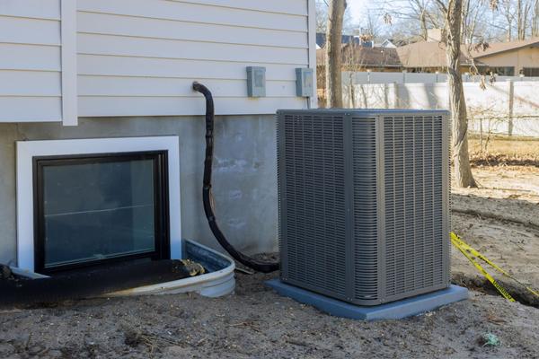 An AC unit located on the ground, near a basement window well that is on the side of a house that has white paneling.