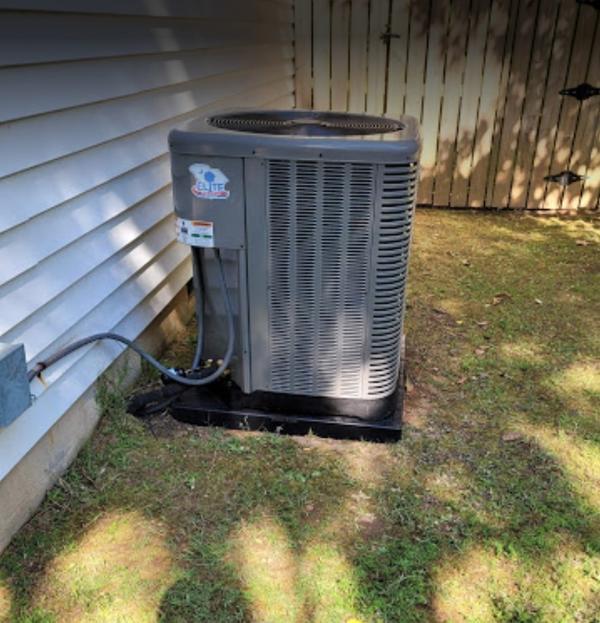 An AC Unit located in a grassy spot on the side of a white paneled house with a wooden gate showing behind the AC unit.