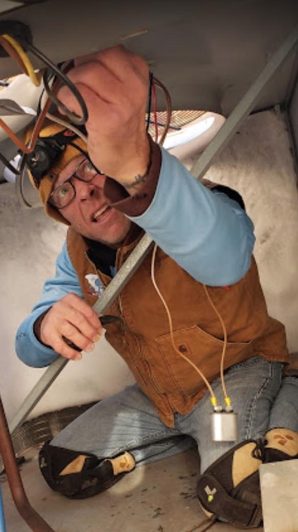 Elite Air Technician wearing glasses, a brown vest, and knee pads, working on a heat pump repair.