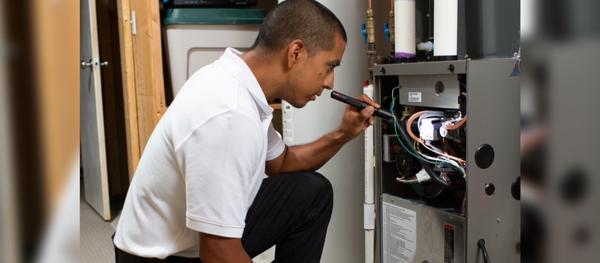 A technician wearing a white collared shirt and black pants is kneeling down in front of a furnace, holding a flashlight and checking things out on the unit.