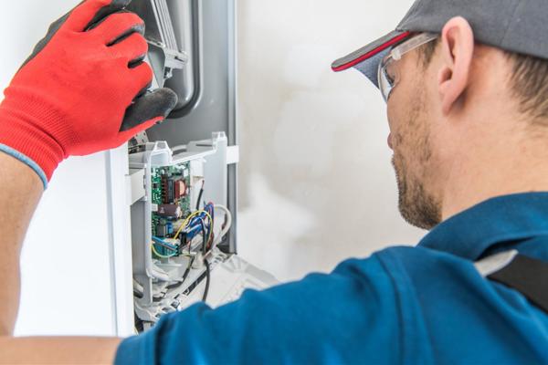 A technician wearing a blue shirt, gray ball cap, clear safety glasses, and a bright red work glove is taking a look at a furnace unit.