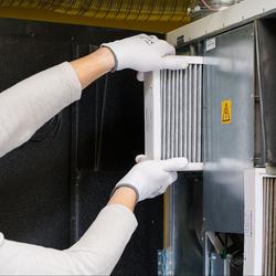 Tech wearing a white hard hat, gloves, long sleeve shirt, and black glasses is pulling out a furnace filter.