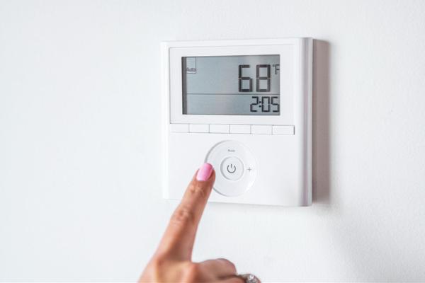 Woman with pink painted nails pointing at a digital thermostat display at 68 degrees, affixed to an all white wall.