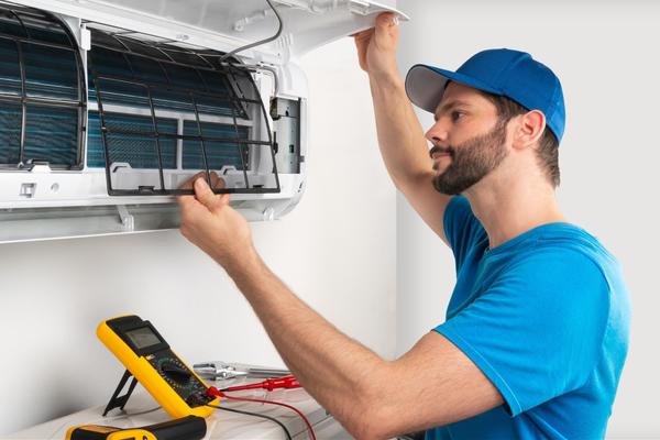 Man wearing a blue shirt and blue baseball cap putting a black mesh filter in a ductless ac unit affixed to a white wall, with various tools placed out in front of him