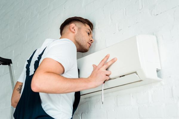 Tech wearing blue overalls and white shirt is inspecting a ductless ac unit with a Phillips-Head screwdriver in his right hand.