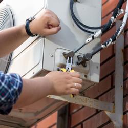 Man wearing a blue, plaid long-sleeve shirt, a watch on his left wrist, and a ring on his right ring finger is using pliers to adjust a part on an AC unit
