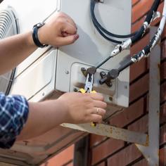 Man wearing a blue, plaid long-sleeve shirt, a watch on his left wrist, and a ring on his right ring finger is using pliers to adjust a part on an AC unit